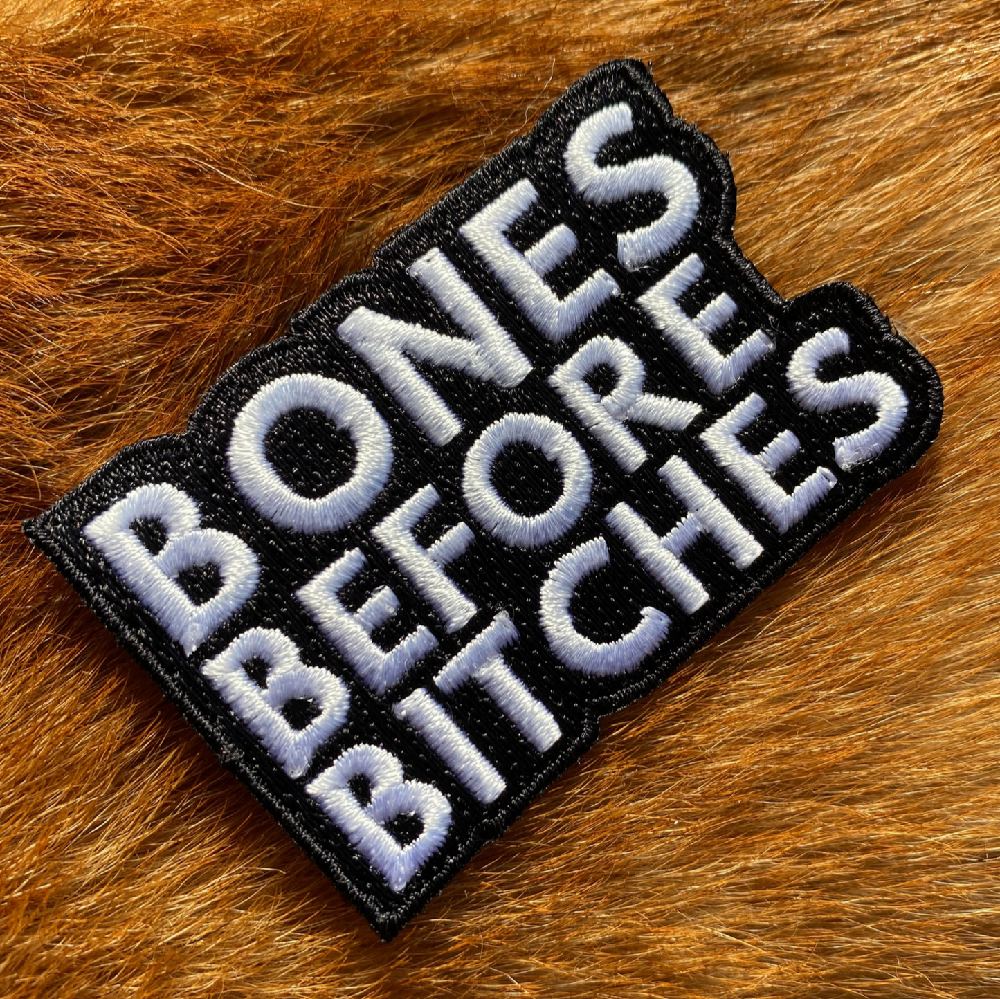Embroidered black and white patch with the text Bones Before Bitches on a background of ginger dog fur.