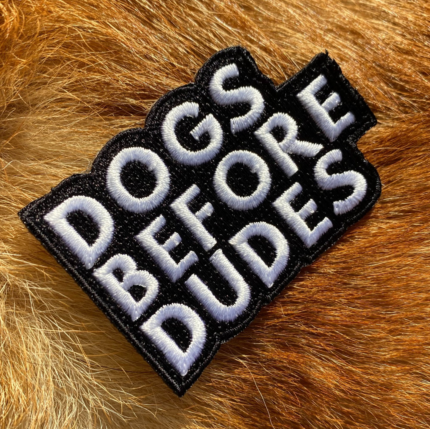 Embroidered black and white patch with the text Dogs Before Dudes on a background of ginger dog fur.