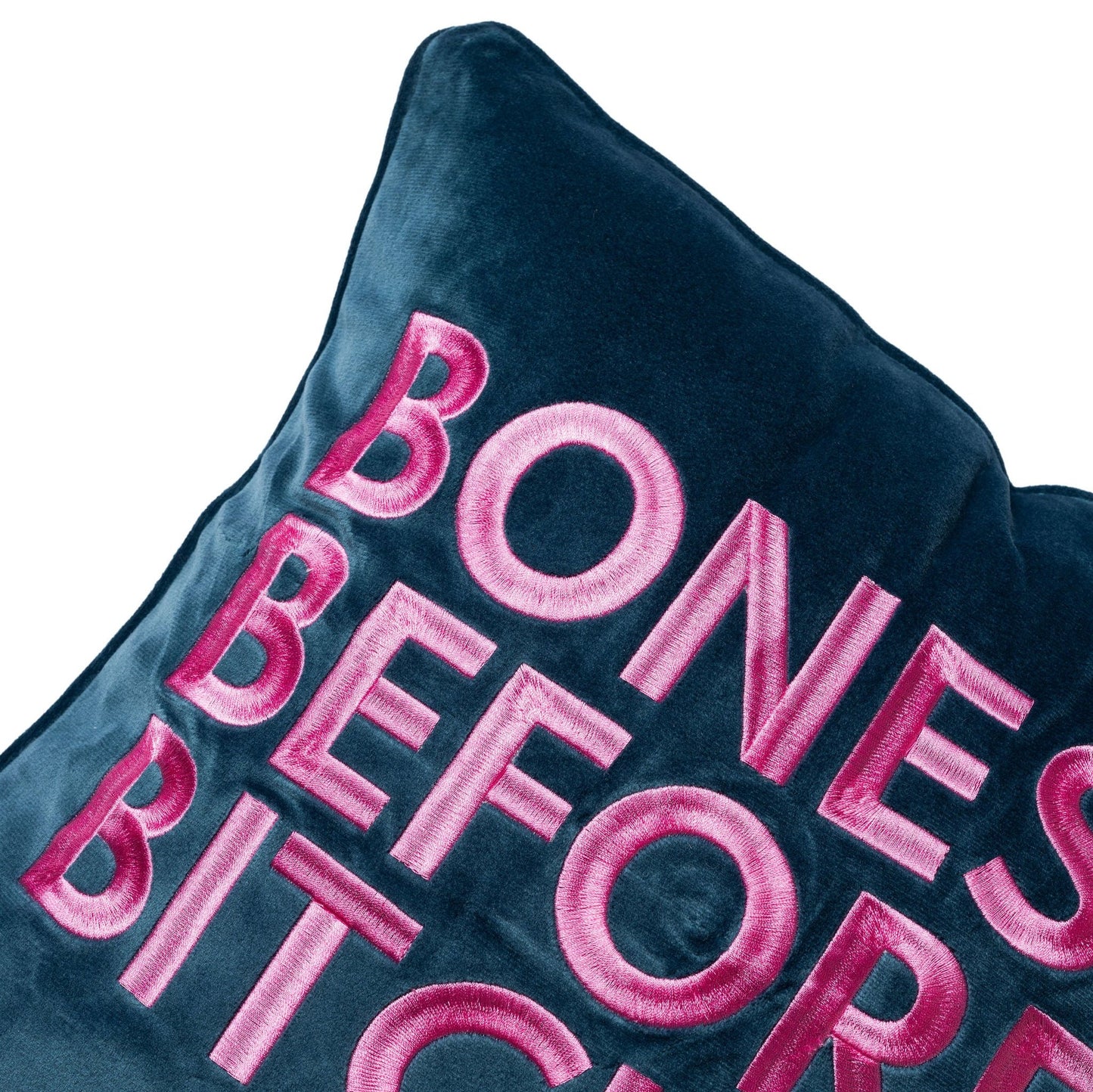 side view of velvet square pillow with Bones Before Bitches embroidered on to it. Peacock base with pink embroidery.